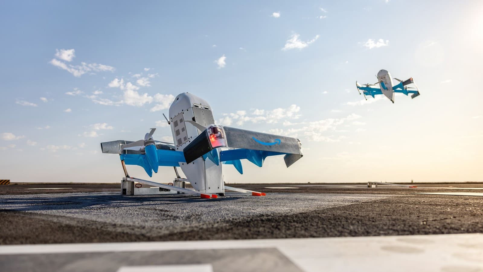 Two Amazon Prime Air MK30 delivery drones, one parked on a launch pad and one flying nearby