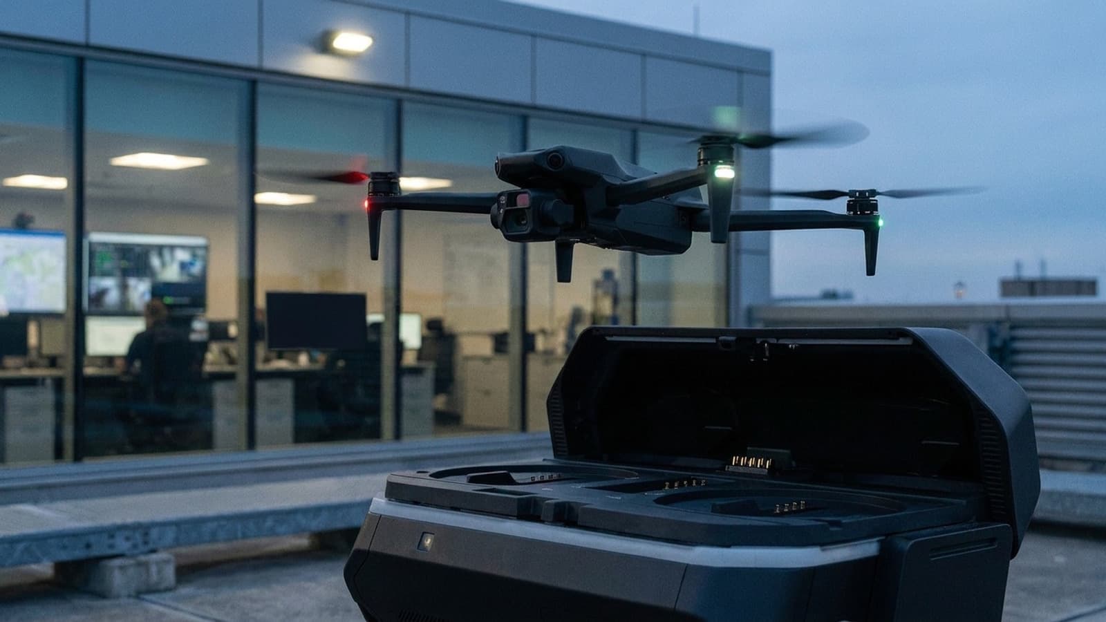 Public-safety drone returning to a rooftop dock outside a glass-walled operations room with live aerial views